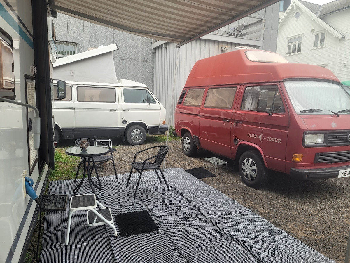 Two VW vans are parked in a gravel area, one red marked as 'Club Joker' and another white. A striped outdoor mat covers the ground, with a black chair and a small table positioned nearby, offering a cozy outdoor seating space.