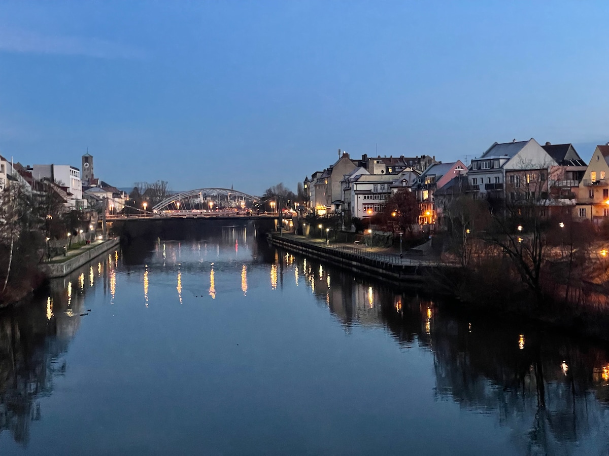 The image captures a tranquil riverside scene at dusk, with softly lit buildings reflecting on the calm water. A bridge arches gracefully in the background, and trees line the riverbank, contributing to the evening's serene ambiance.