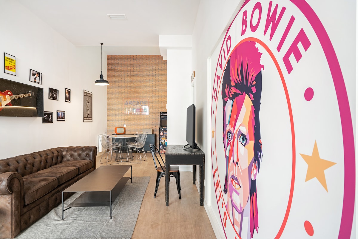 A spacious living area features a brown Chester sofa and a sleek coffee table. A vibrant mural of David Bowie adds a striking focal point. A dining table with transparent chairs is visible in the background, along with decorative wall art that reflects a musical theme.
