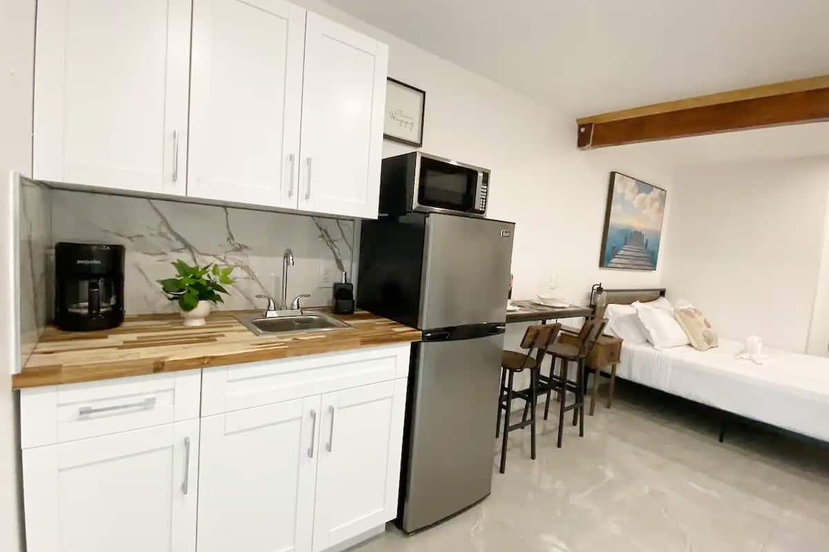 Rectangular room with shiny grey tile flooring. The room features a kitchenette with a sink and coffee maker. There is a small refrigerator and a rectangular high top table with two stools. There is a queen size bed in the background, with a painting of a dock above it. 