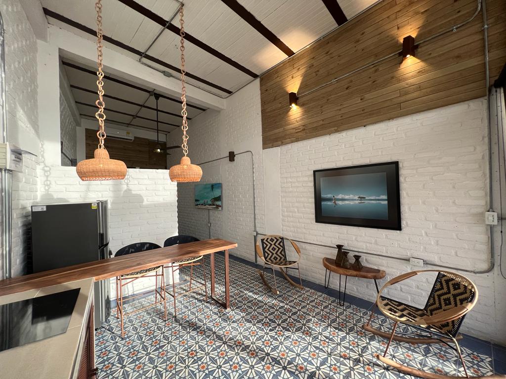 A well-lit dining area features a wooden table complemented by two woven chairs. The floor showcases intricate tile patterns, while the walls consist of exposed brick and wood accents. Two pendant lights illuminate the space, and a refrigerator is visible along the wall.