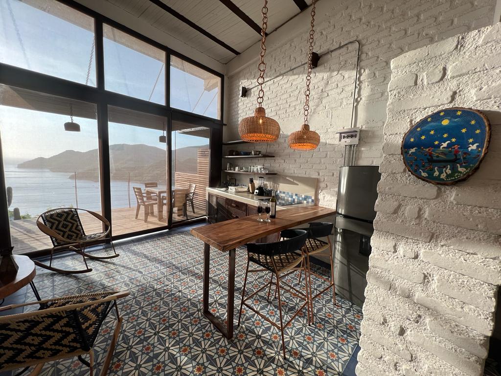 A stylish kitchen area features a wooden table with chairs, surrounded by large glass doors that provide a view of the bay. Rattan light fixtures hang above, and decorative tiles line the floor, enhancing the spacious and bright atmosphere.