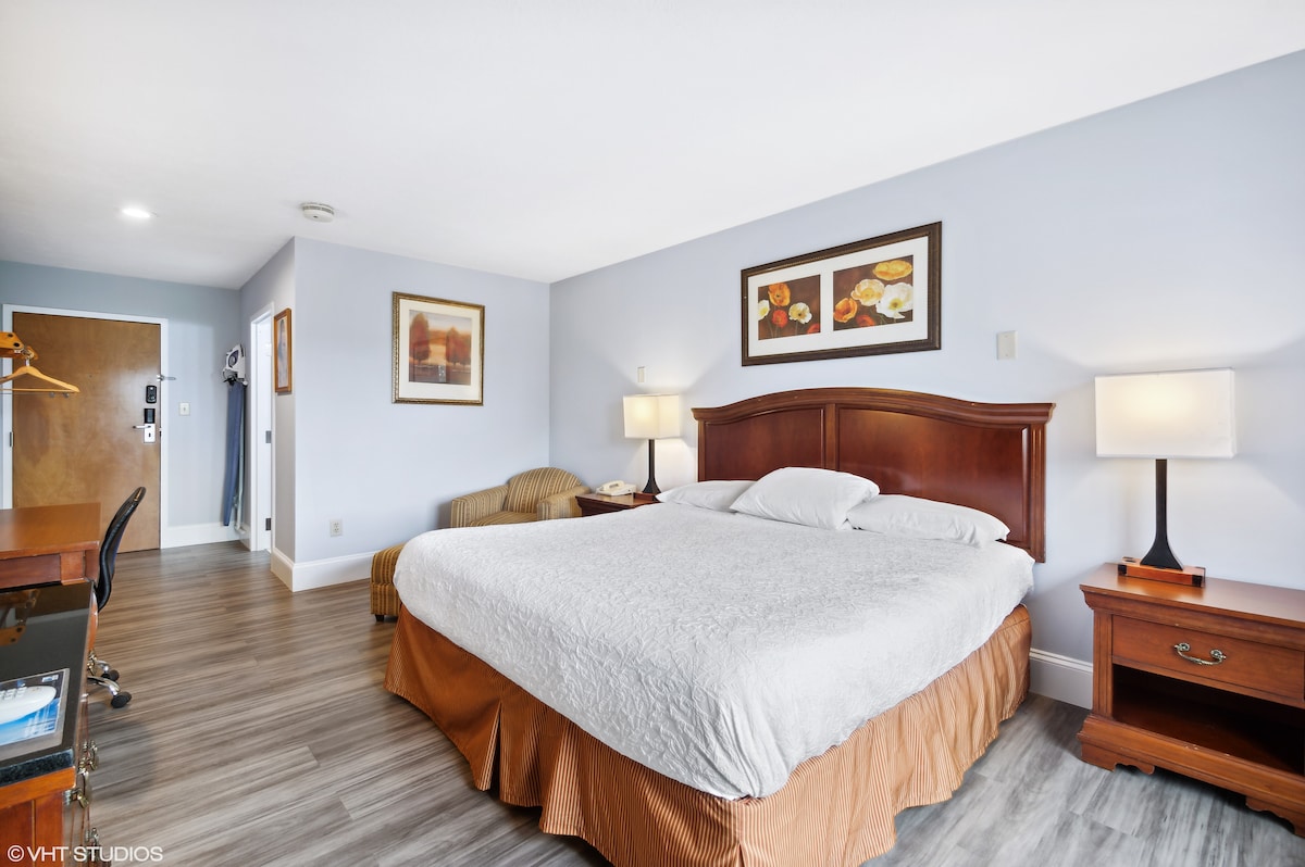 A spacious hotel room features a large bed with light bedding and an orange skirt. A seating area with a soft chair is visible, along with a wooden desk and dresser. Soft light is provided by bedside lamps, and artwork adorns the walls.
