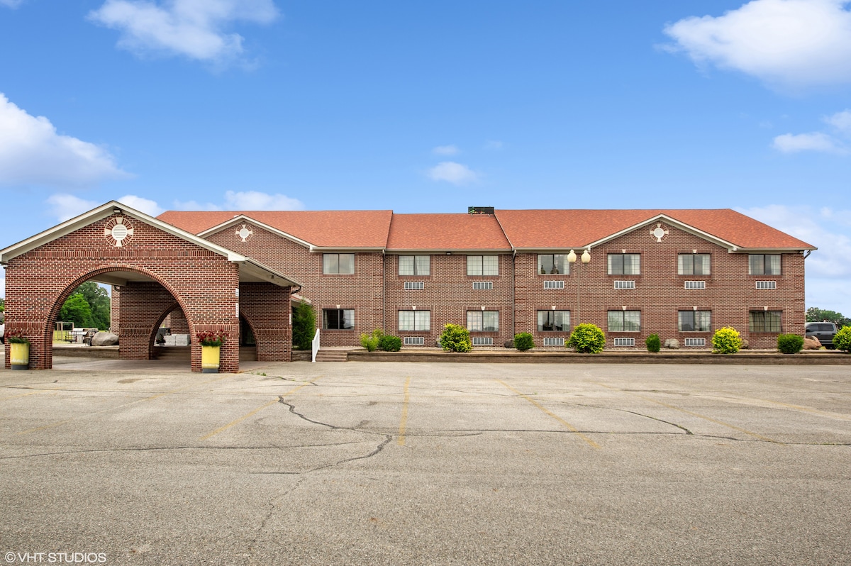 The exterior of the hotel is shown, featuring a two-story brick building with a red-tiled roof. A covered entrance is visible, along with ample parking space in front. Neatly trimmed shrubs and trees enhance the landscaping.
