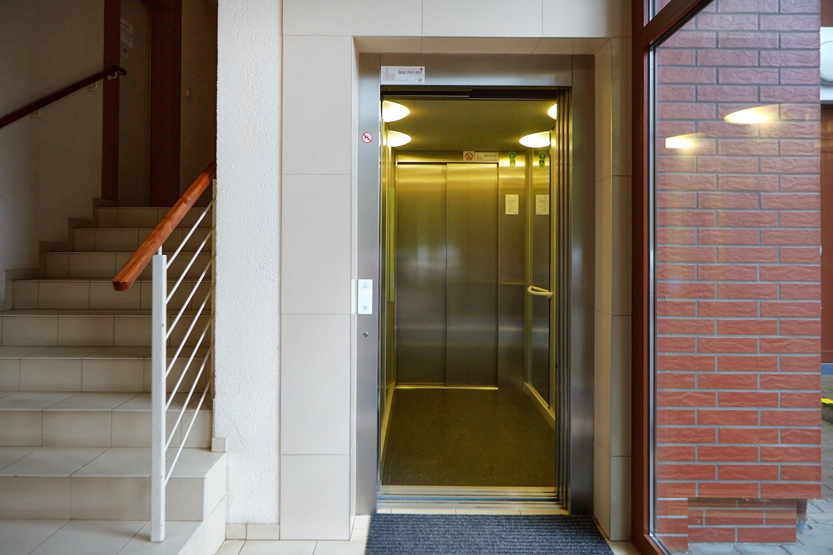 A modern elevator entrance is visible, with sliding doors that open to a spacious interior. Adjacent stairs are framed by a wooden railing, and light enters through large windows, providing a bright, welcoming atmosphere.