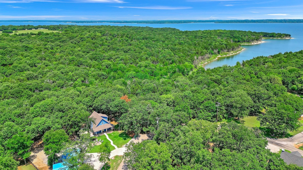 An aerial view captures the spacious property surrounded by lush greenery and trees, with the nearby Lake Texoma visible in the background. The house sits within a natural setting, offering a serene escape. The landscape leads towards a tranquil water edge.