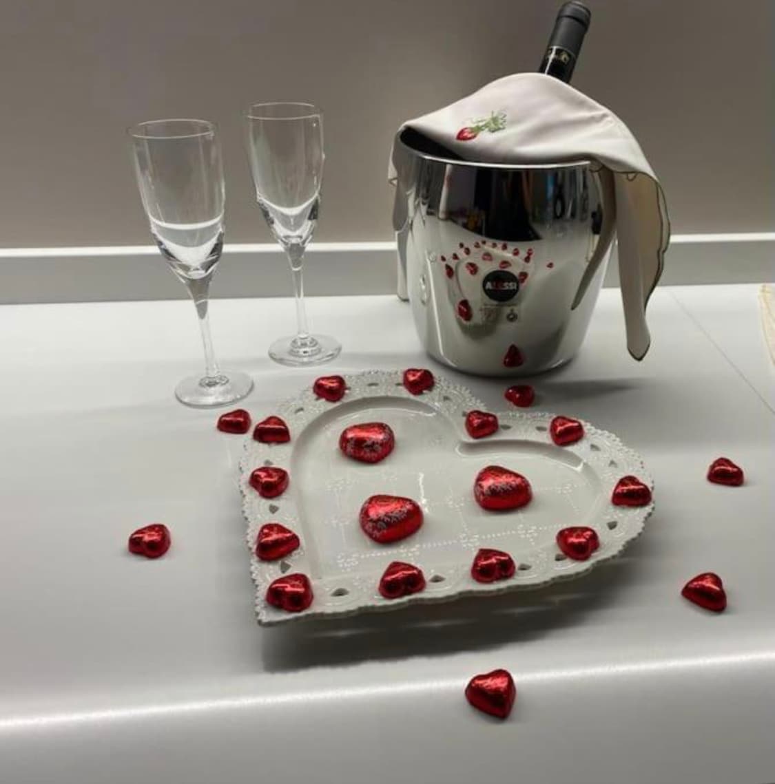 A heart-shaped platter adorned with small red decorative hearts is positioned on a smooth surface. Two elegant champagne flutes are placed nearby, accompanied by a polished silver ice bucket, suggesting a romantic setting for guests.
