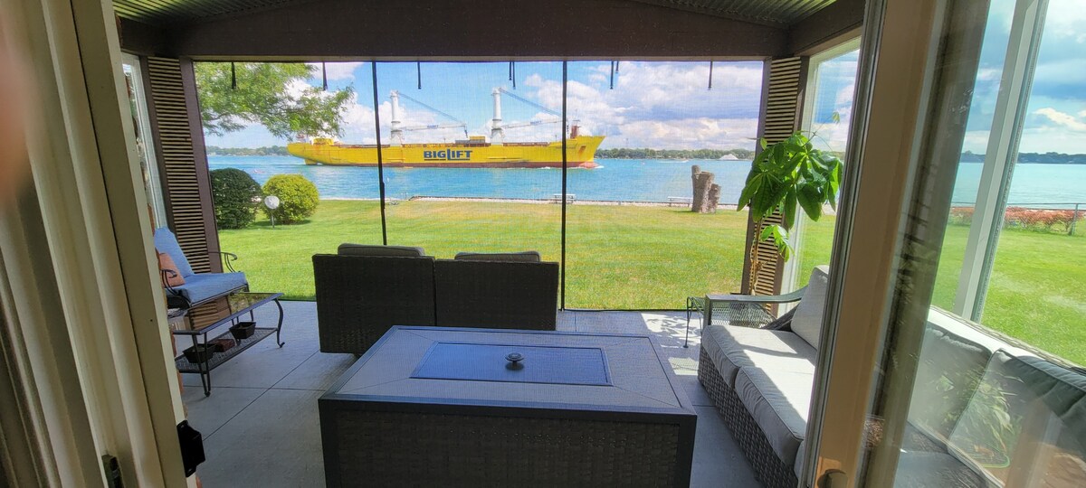 A covered porch features comfortable seating arranged around a central table, with expansive windows providing views of the St. Clair River. A large freighter is seen traveling down the river, with grassy areas and trees visible outside.