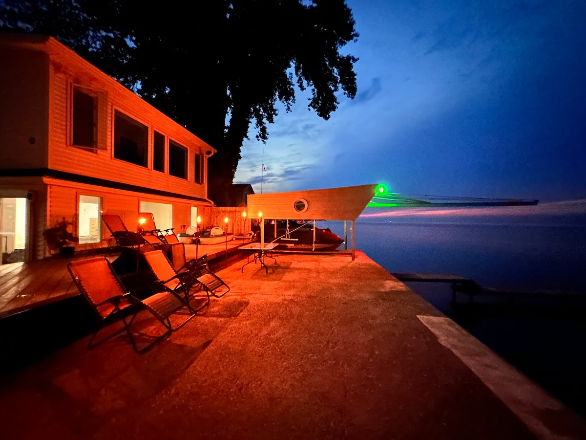 The outdoor deck features several lounge chairs positioned facing the tranquil waters of Lake Ontario, illuminated by subtle evening lights. A clearly visible boat dock extends out towards the lake, complementing the peaceful evening sky.