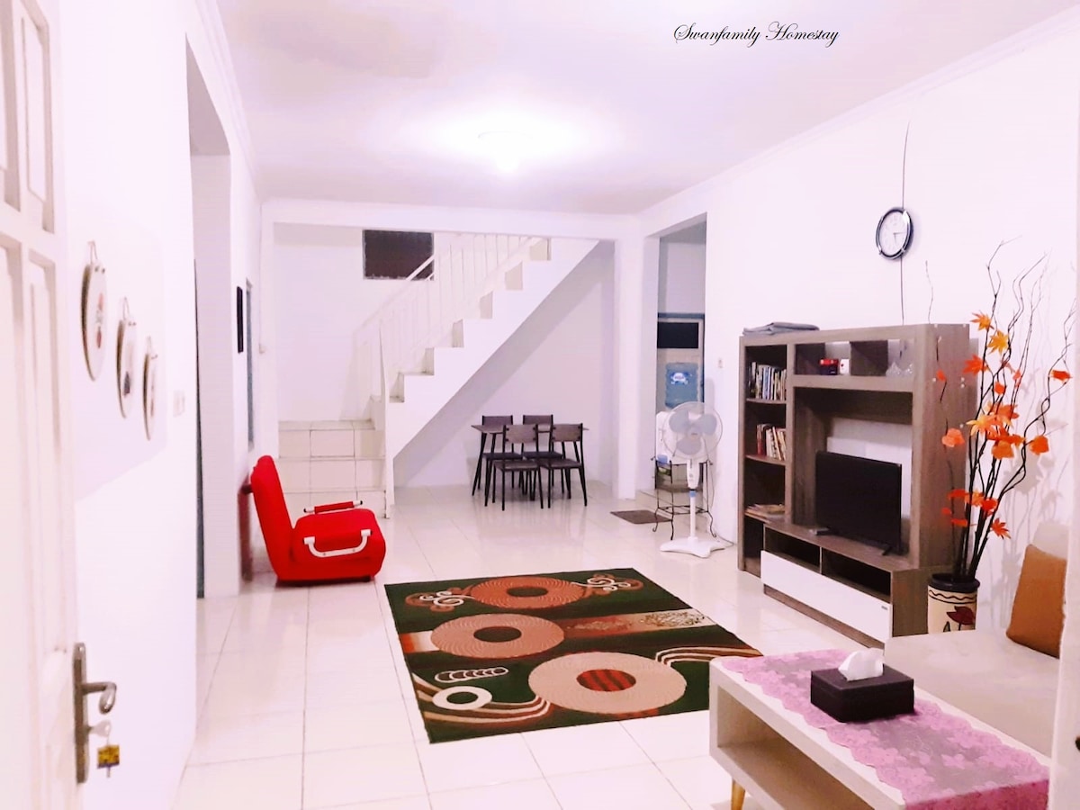 An open living area is presented, featuring a vibrant area rug at its center. A red lounge chair is positioned nearby, along with a wooden dining set accommodating six. A TV unit with a television is visible, complemented by decorative plants and a wall clock.