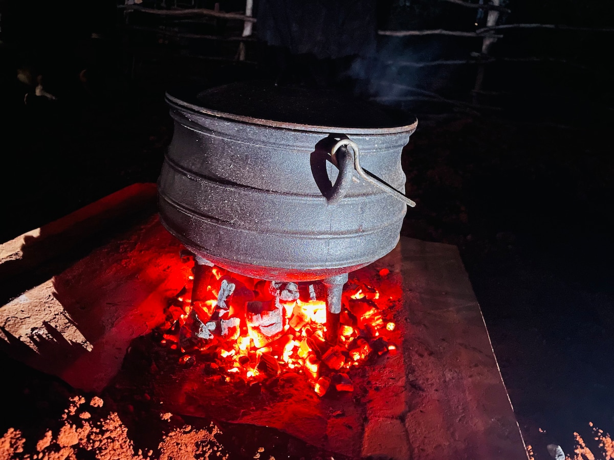 A cast iron pot is placed over glowing embers, with heat rising and creating a soft haze in the air. The pot's dark surface contrasts with the vibrant red and orange hues of the coals, illuminating the surrounding area in the evening tranquility.