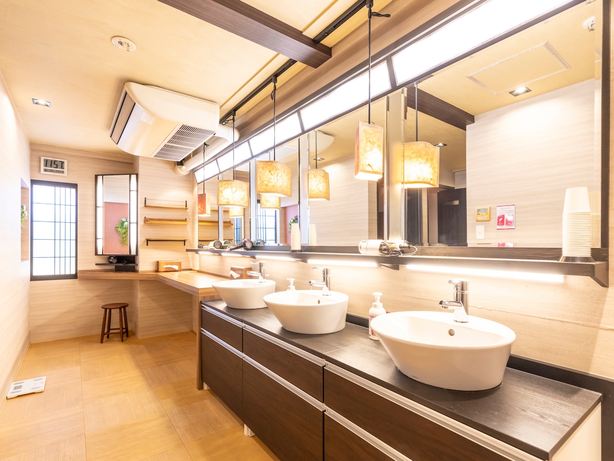 A modern bathroom features dual vessel sinks positioned on a sleek dark wood vanity. A large illuminated mirror enhances the space, complemented by stylish pendant lighting. Natural light filters through a window, and a wooden stool is visible against the wall.