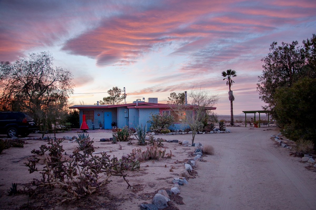 A desert homestead is set against a vibrant sunset sky, with soft pastel clouds overhead. The property is surrounded by native desert plants and a winding dirt path leading to the entrance, evoking a sense of tranquility and seclusion.