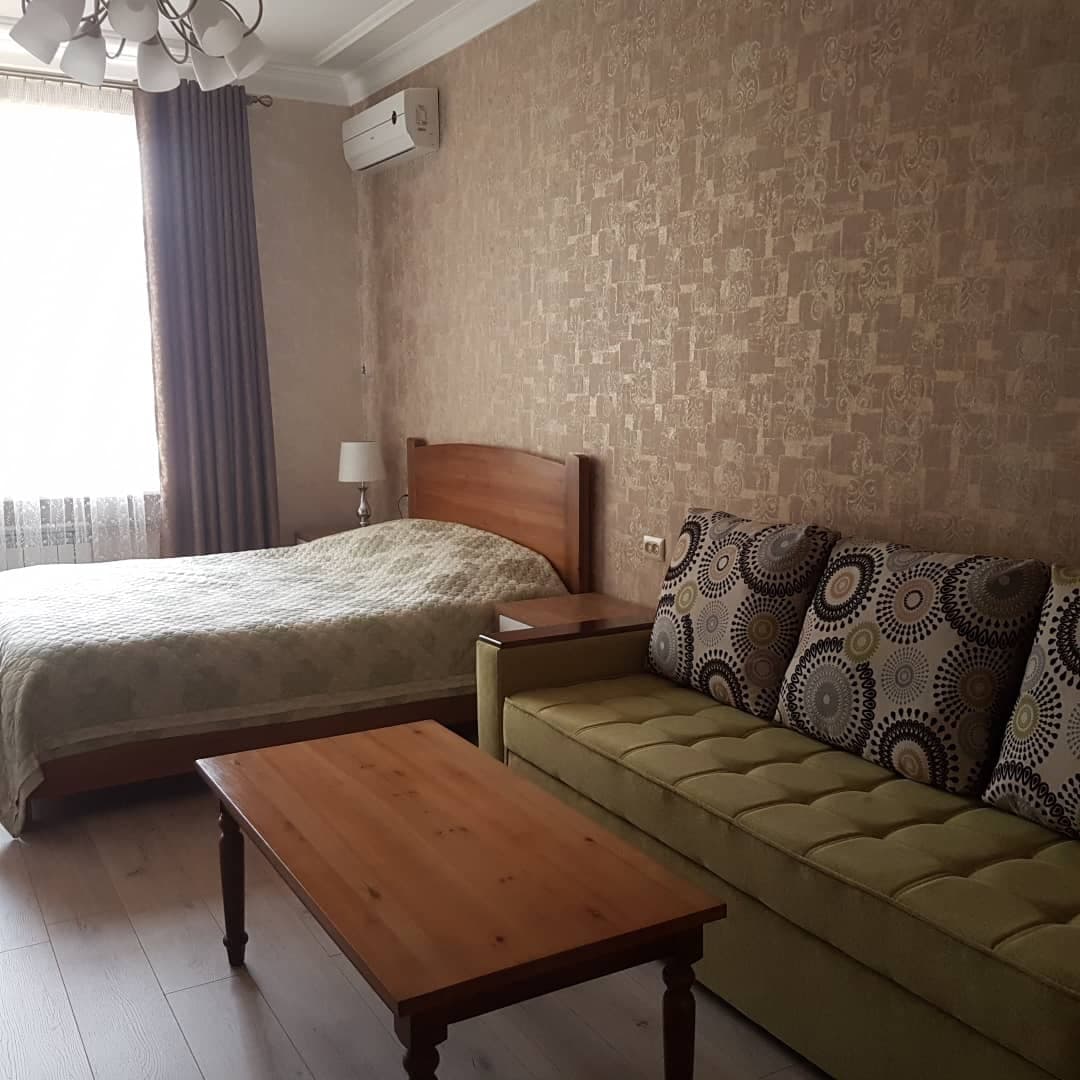 A well-appointed living space features a spacious bed adorned with a light-colored covering and a patterned sofa beside a wooden coffee table. Natural light filters in through a large window draped with sheer curtains, providing a cozy atmosphere.