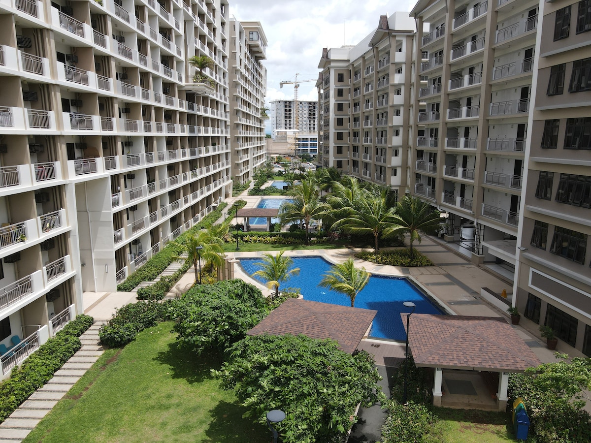 A view of the landscaped courtyard features multiple swimming pools surrounded by palm trees and shaded cabanas. The buildings on either side provide a sense of enclosure, with walkways leading through the greenery. Natural light enhances the inviting atmosphere of the outdoor space.