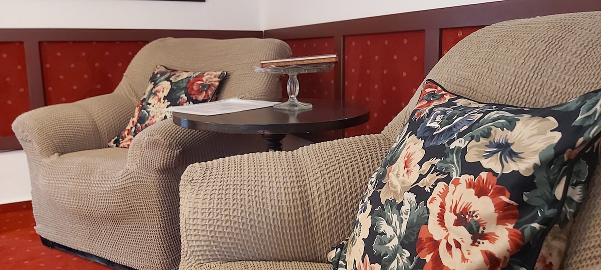 Two comfortable armchairs are placed beside a small wooden table, which holds a decorative dish. The chairs feature floral-patterned cushions and are set against a backdrop of textured red wall panels.