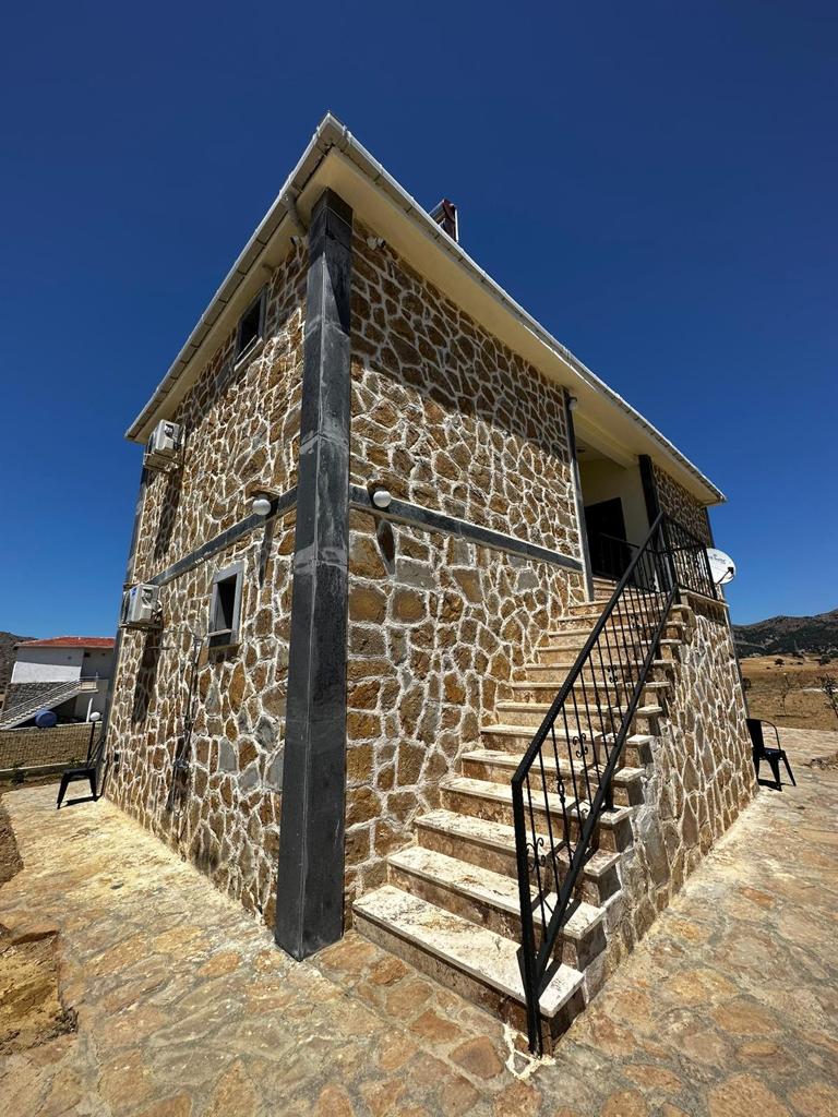 The exterior of the stone house is showcased, highlighting the textured facade and the staircase leading to the entrance. Black chairs are positioned nearby on a stone pathway, with a clear blue sky above emphasizing the building's charm in its natural surroundings.