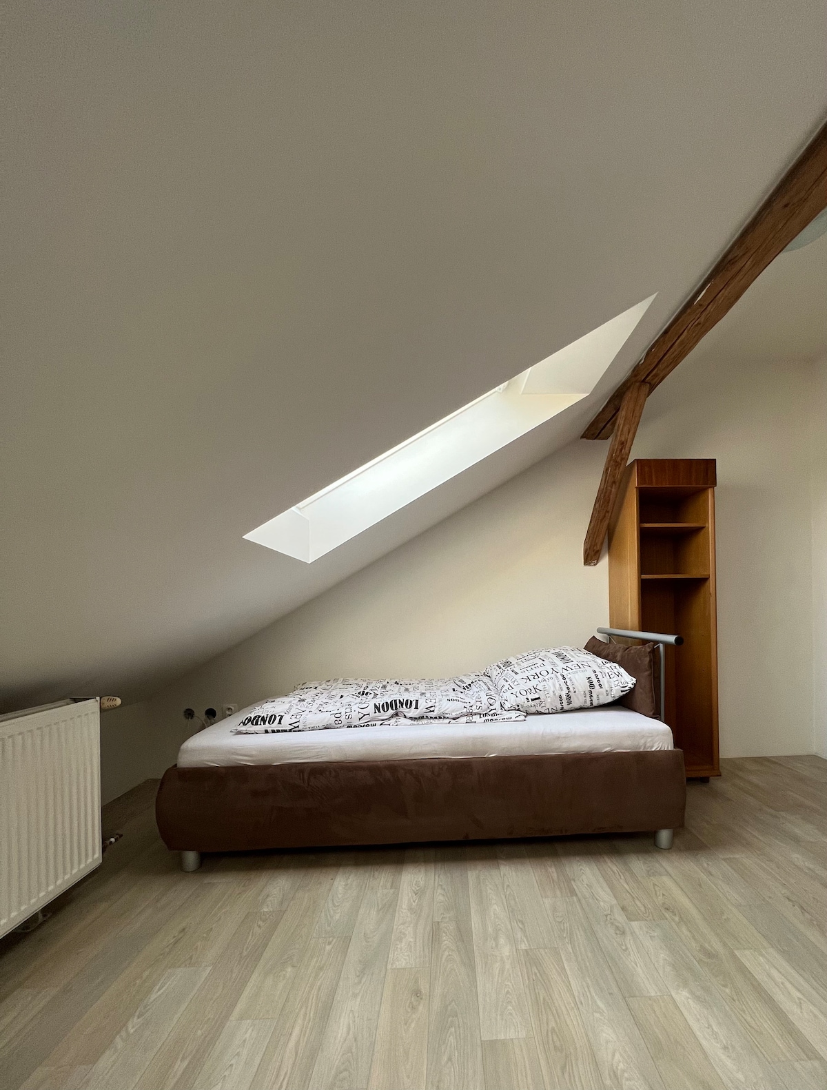 A cozy bedroom features a spacious bed with a neutral-toned duvet positioned against a sloped ceiling. A skylight allows natural light to fill the room, and a tall wooden shelving unit stands nearby, providing additional storage. Light wood flooring completes the warm atmosphere.
