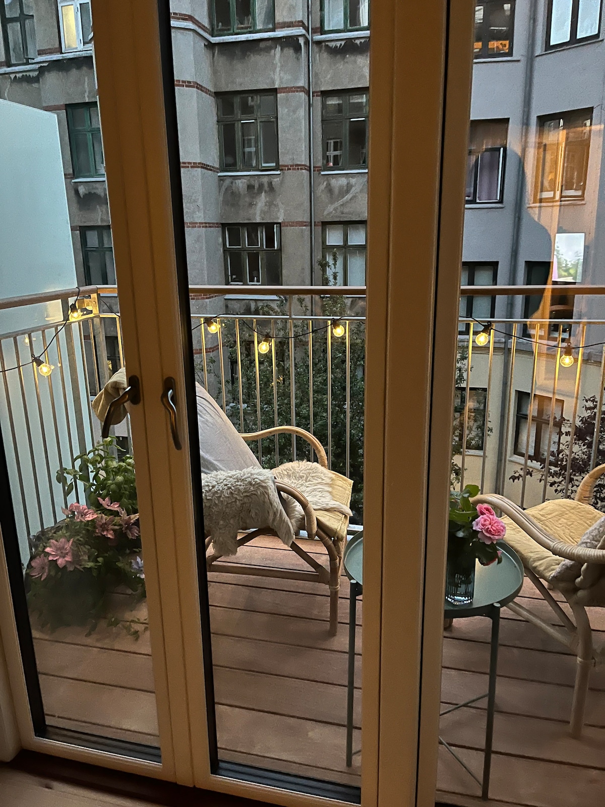 A balcony is visible through glass doors, featuring two rattan chairs and a small round table. Soft lighting from decorative string lights enhances the space. Potted plants with vibrant blooms are placed strategically, contributing to a tranquil outdoor setting that connects to the city view.