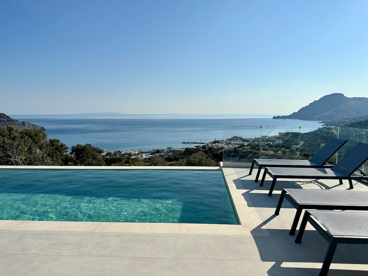 A private pool reflects the clear blue skies and stunning panoramic views of the Plakias coastline. Several loungers are positioned nearby, inviting relaxation and sunbathing. The tranquil waters of the pool seamlessly blend into the expansive seaview, enhancing the serene outdoor ambiance.