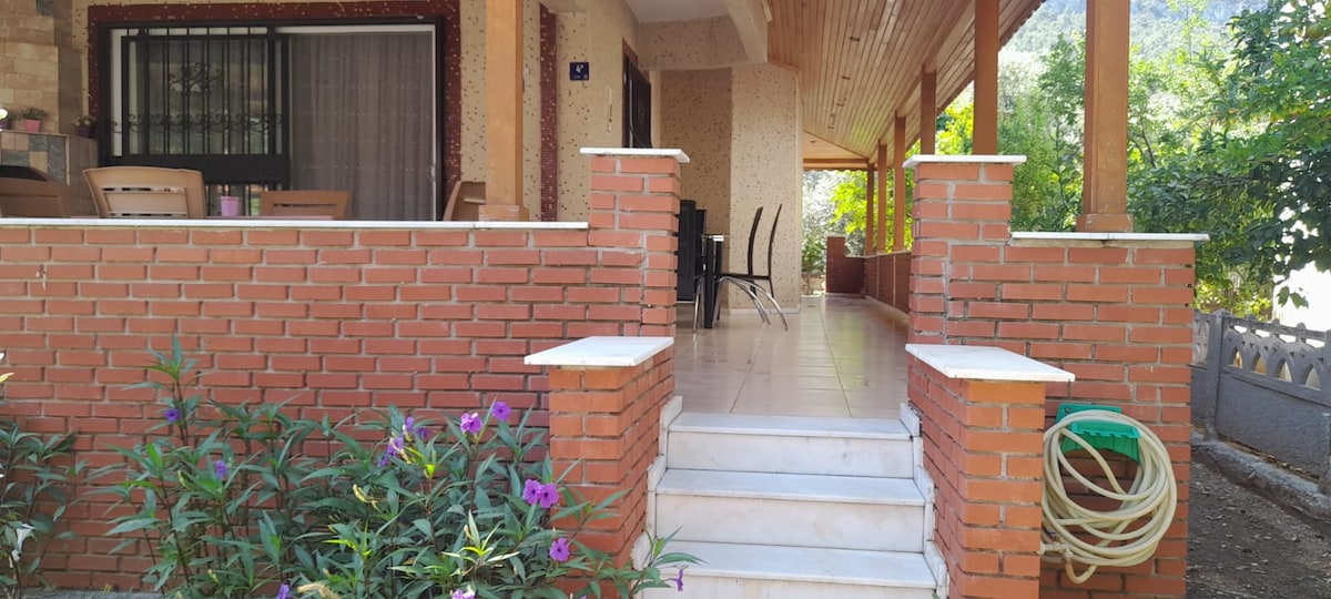 A spacious porch is viewed from the steps, featuring a tiled surface and brick railings. A dining table with black chairs is visible under the shaded area. Lush green plants and vibrant flowers are situated at the base of the steps, enhancing the outdoor setting.