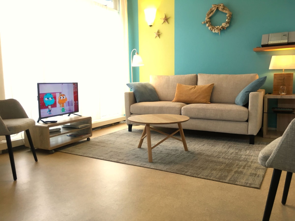 The living area features a light-colored sofa with cushions and a decorative throw pillow, facing a television. A wooden coffee table rests on a soft area rug. Warm lighting is provided by lamps, while colorful walls add character to the space.