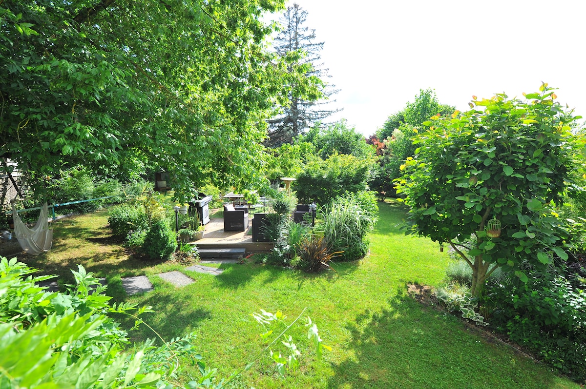 An expansive garden is surrounded by lush greenery, with various plants and shrubs creating a natural atmosphere. A wooden deck area is visible, equipped with seating, while a hammock is positioned among the trees, offering a relaxing spot.