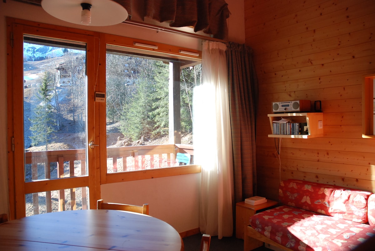 A warm living area is depicted, featuring large windows with wooden frames that provide ample natural light. A round wooden table is visible, accompanied by a red cushioned sofa with patterned fabric. Shelves with a radio and books are mounted on the wooden wall.