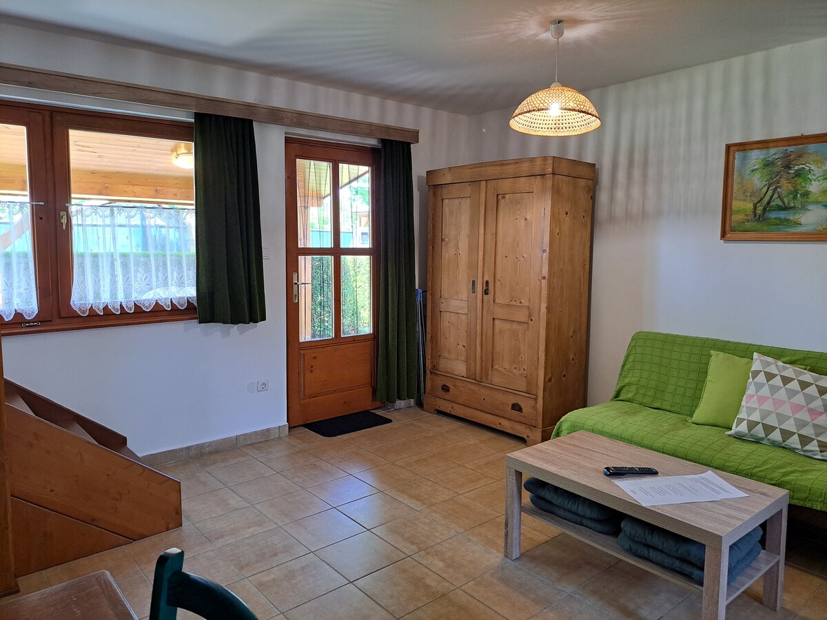 A welcoming living area features large windows allowing natural light to fill the space. A green sofa sits beside a wooden coffee table, with a decorative cushion adding color. A wooden wardrobe and a simple wall art piece enhance the cozy environment.