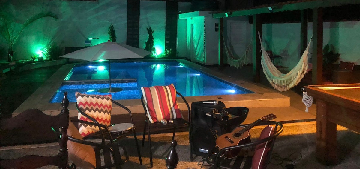 Seating options are arranged around the pool area, featuring a mix of colorful chairs and cushions. A suspended hammock is visible, alongside soft lighting that enhances the serene ambiance. The pool, illuminated in blue, serves as a central focal point in the outdoor space.