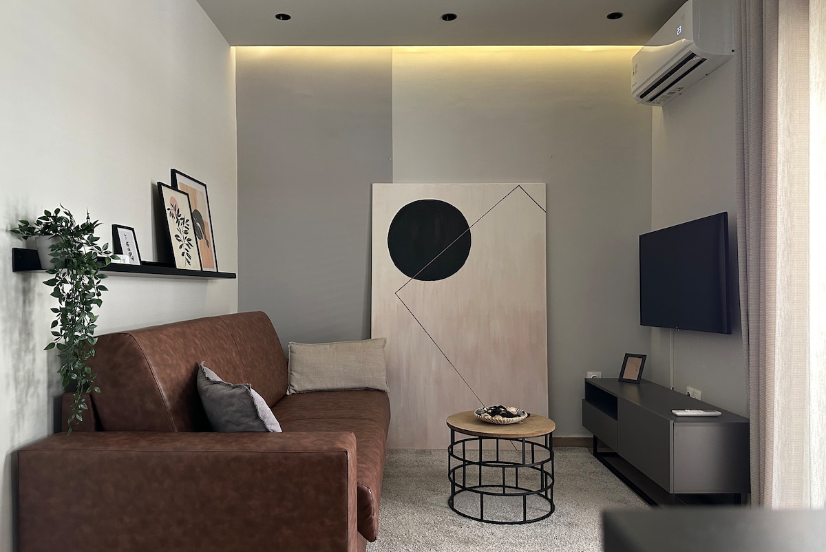 A cozy living area is showcased with a brown sofa against a light grey wall. A modern piece of wall art is displayed beside a television on a low unit. A round coffee table sits in front, with decorative items arranged atop soft carpeting.