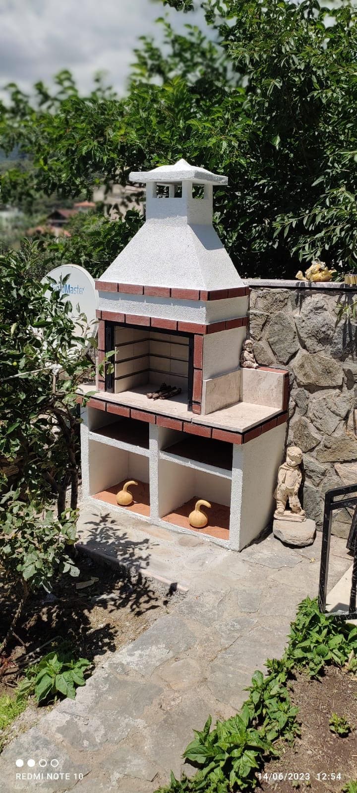 A decorative outdoor barbecue grill is positioned within a stone-fenced garden area. The grill features a white and red tiled design with two storage compartments below. Surrounding the grill are various plants and statues, contributing to a serene garden ambiance.