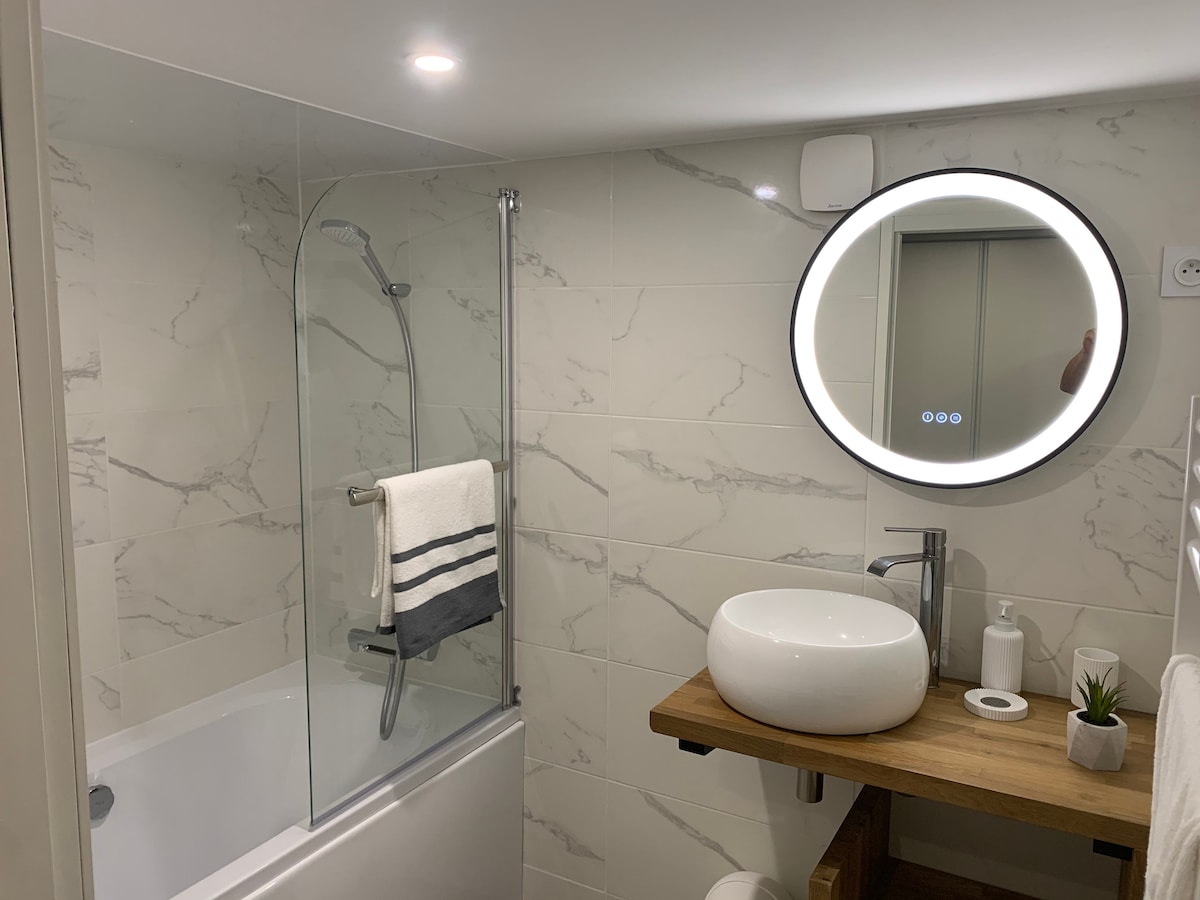 A modern bathroom is presented with a sleek, round mirror illuminated by built-in lighting. A wooden vanity supports a white vessel sink, while a glass shower enclosure showcases a handheld showerhead. Towels are neatly displayed, and natural light is enhanced by light-colored marble tiles.