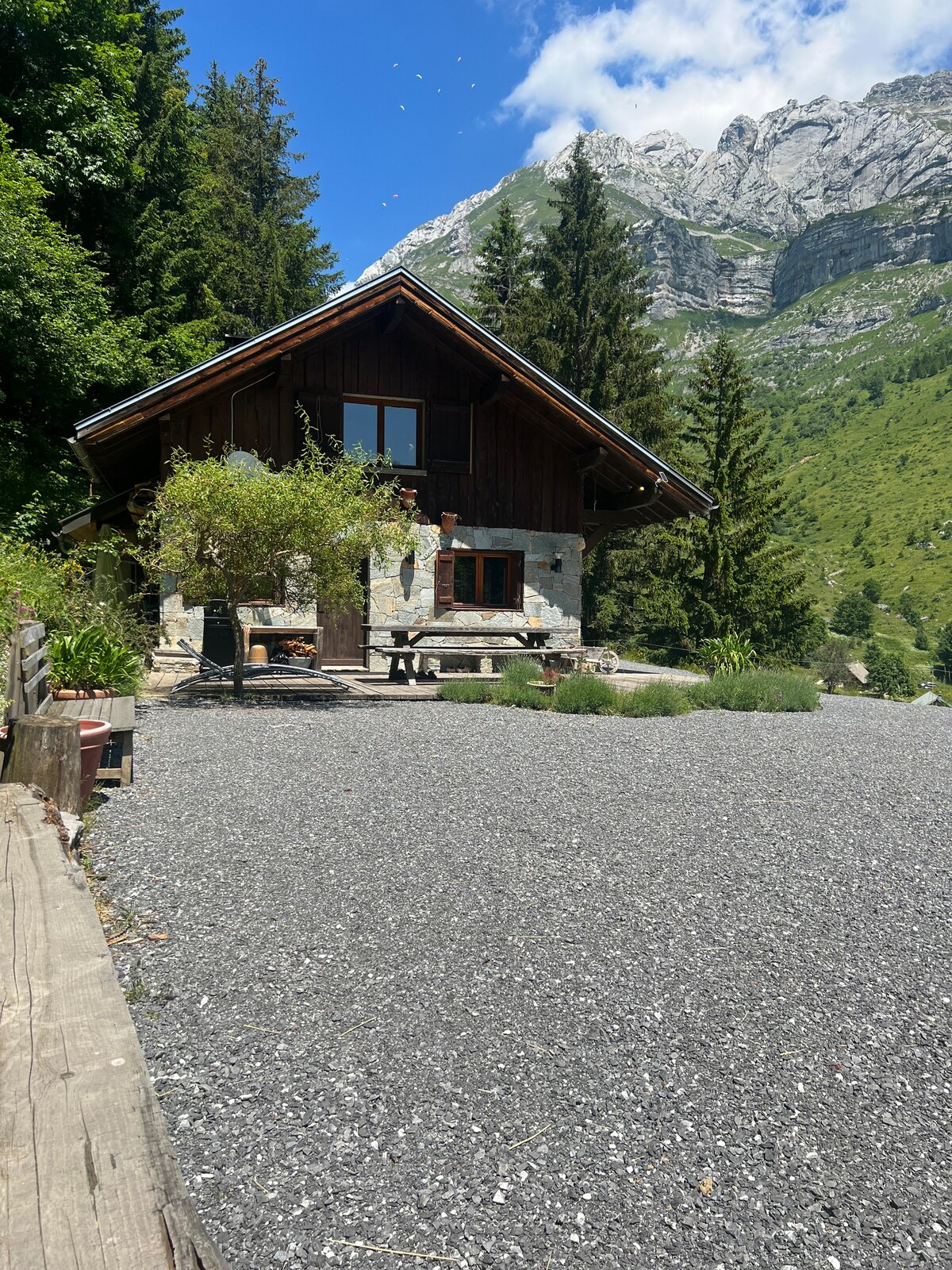 A wooden chalet is surrounded by lush greenery and mountains, presenting a harmonious blend of natural elements. A stone terrace features a picnic table and a grill, with gravel leading to the entrance, inviting outdoor gatherings amidst the scenic landscape.