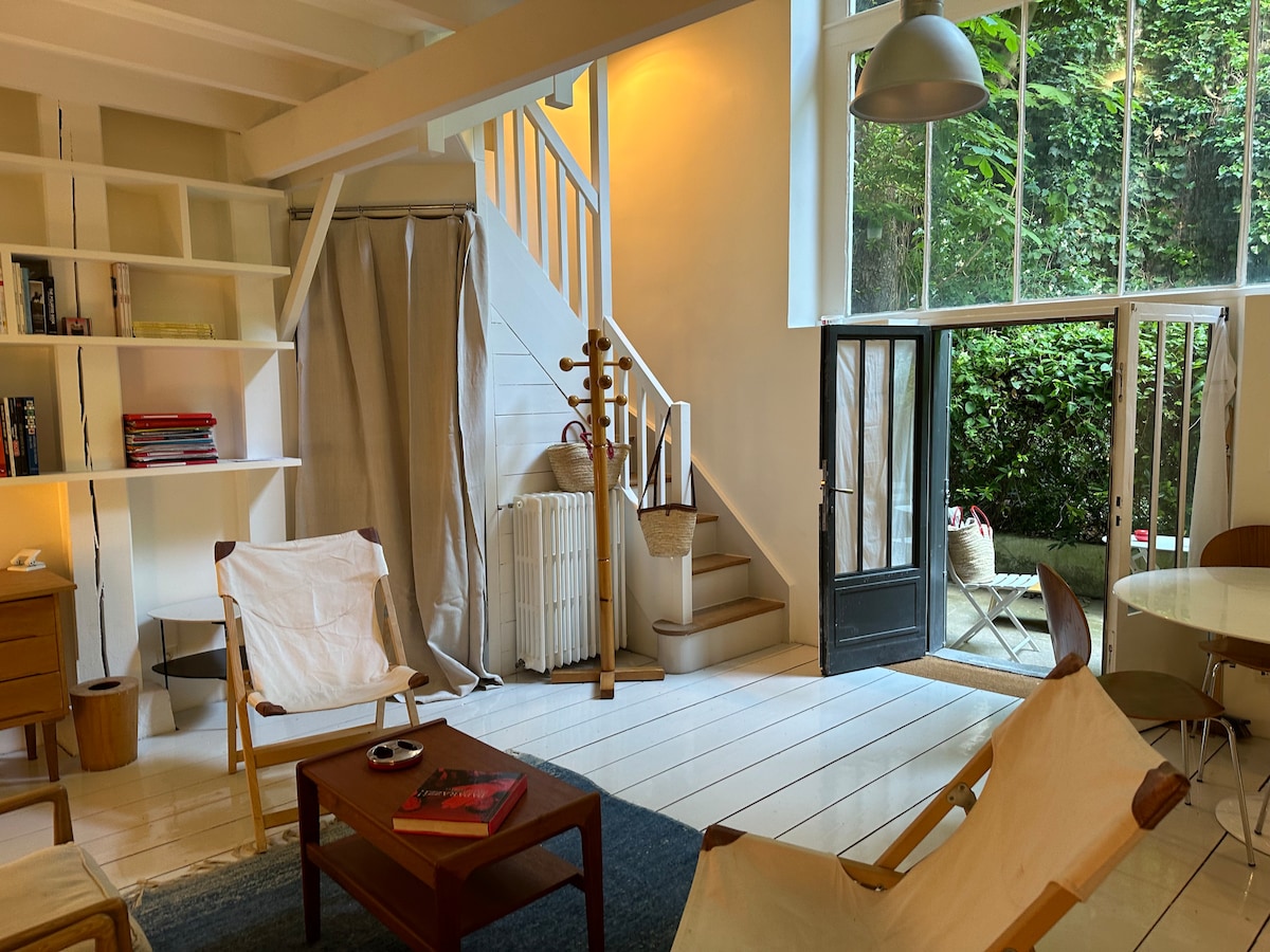 A spacious living area features a large glass window providing views of lush greenery. A light rug anchors seating with wooden chairs, and a round dining table is set nearby. A staircase leads to the mezzanine, while shelves display books and decorative items.