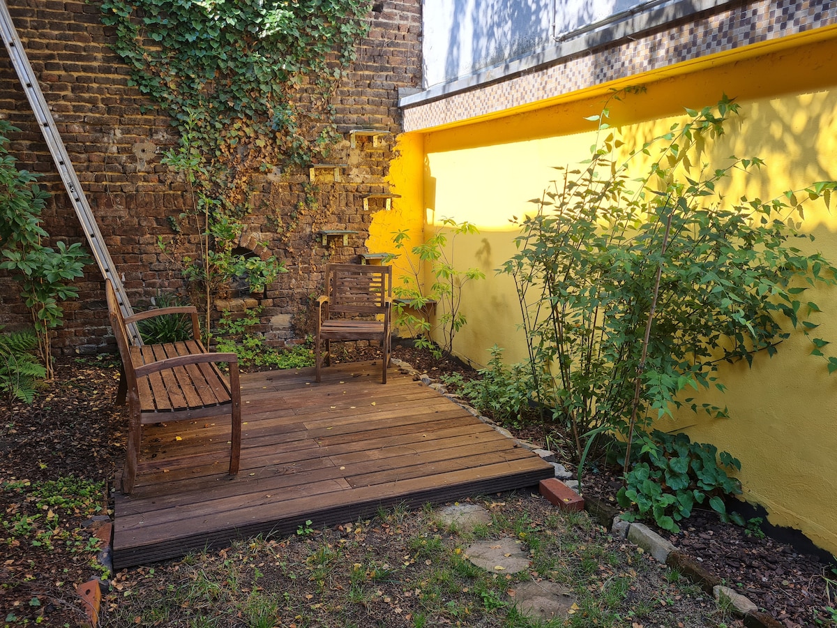 A small outdoor area is showcased, featuring two wooden chairs positioned on a raised wooden deck. Surrounding greenery includes bushes and small plants, complemented by a backdrop of textured brick walls and a vibrant yellow wall.