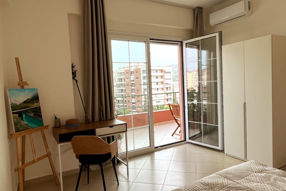 A cozy bedroom is highlighted by large glass doors that open to a balcony. A simple desk and chair are positioned next to an easel with artwork. A spacious wardrobe stands against the wall, while natural light fills the room through the doors.