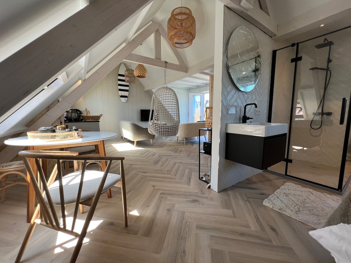 The open living space features wooden flooring and a modern design. A round dining table with wooden chairs sits beside a cozy seating area. A chic, well-equipped kitchenette is visible, along with a spacious bathroom, showcasing a large mirror and a stylish shower.