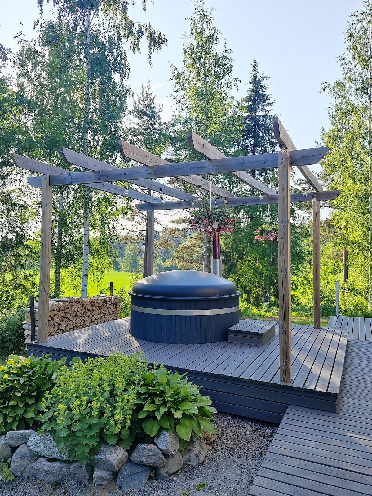 An outdoor deck features a black hot tub beneath a wooden pergola adorned with hanging plants. Surrounding greenery adds to the serene atmosphere, and a neatly stacked pile of firewood is visible nearby.