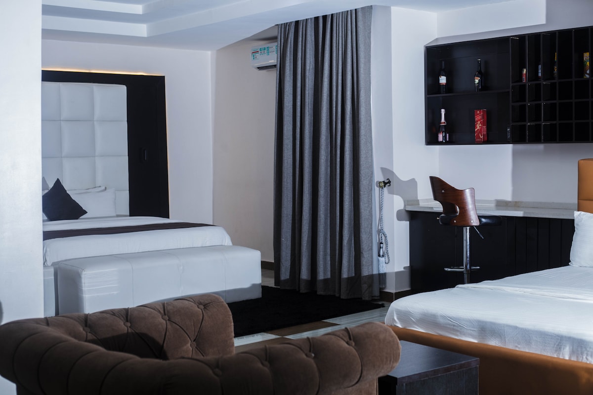 The spacious room is divided into separate areas featuring a double bed with a plush headboard and a second bed. A modern workspace and a cozy sofa sit adjacent to the in-room bar. Soft curtains frame the windows, enhancing the comfortable ambiance.