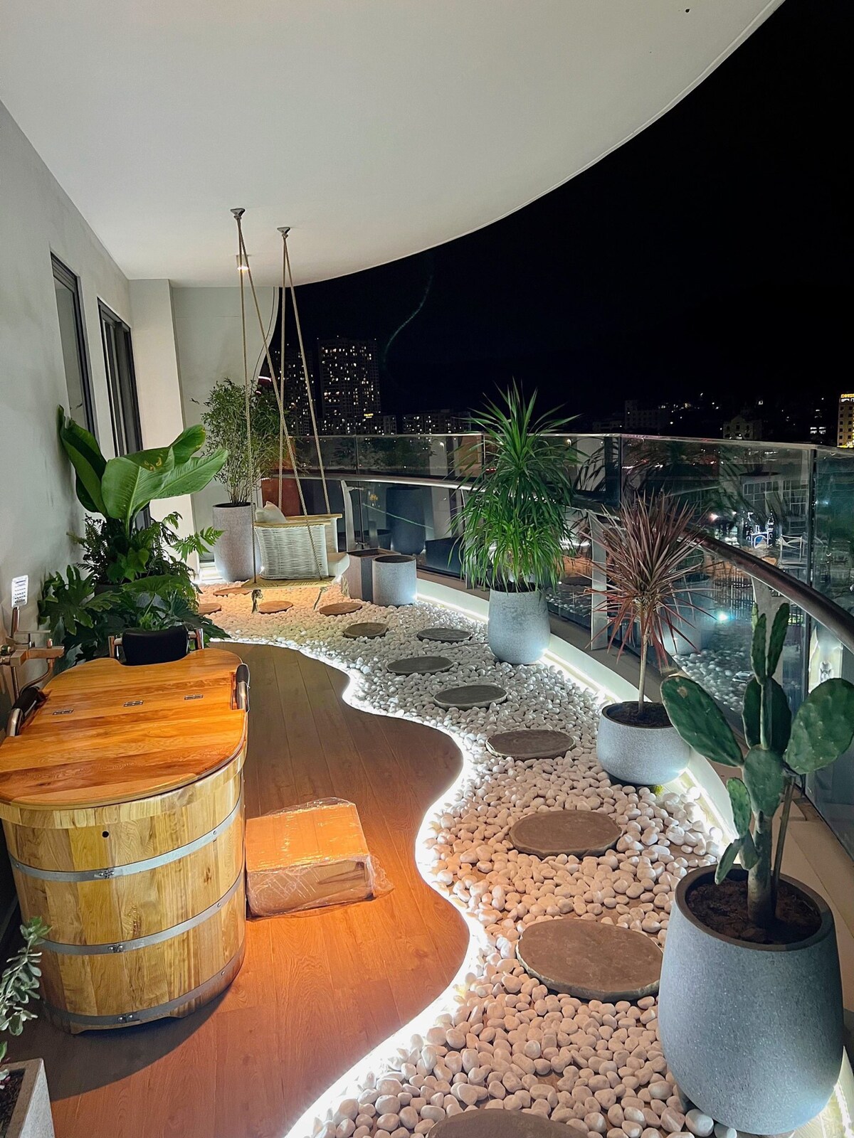 A beautifully designed balcony showcases a winding path lined with smooth stones and illuminated by soft lighting. Potted plants are positioned throughout the space, adding greenery. A wooden barrel serves as a rustic table, complemented by hanging chairs that offer a cozy seating option.