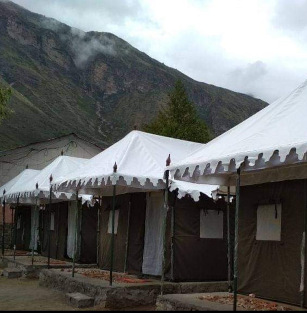 A series of tent accommodations are set against a backdrop of rolling mountains and overcast skies. Each tent features a sloped white roof and dark brown canvas walls, with small windows that provide ventilation. Surrounding greenery adds to the natural ambiance.