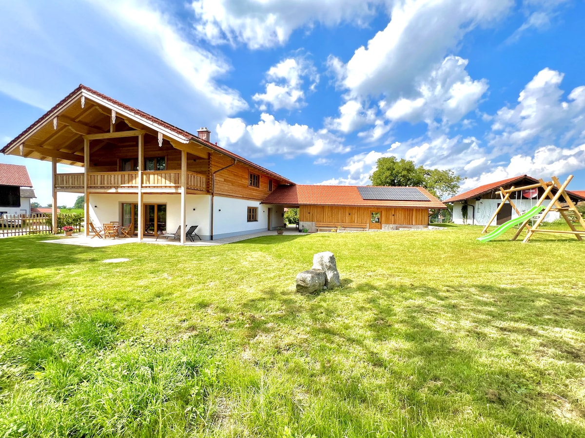 The exterior of a spacious holiday home is shown, featuring a two-story structure with a covered balcony. A well-maintained lawn surrounds the building, complemented by trees and a play structure with a slide. Bright clouds stretch across a blue sky.