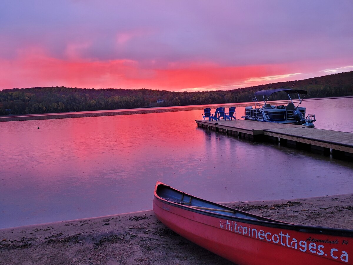 A serene lakeside scene is captured at sunset, featuring a vivid sky painted in shades of pink and purple. A red canoe rests on the sandy shore, while a wooden dock extends into the water, fitted with blue chairs overlooking the tranquil lake.