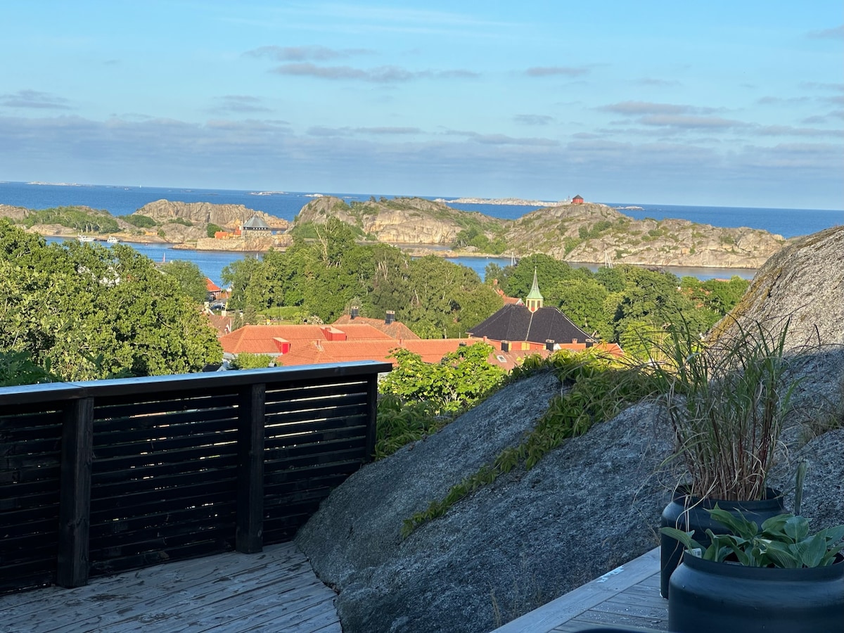 A scenic view of the coastline is captured from a wooden terrace, showcasing the surrounding islands amidst a calm sea. Green trees and red-roofed houses are visible in the foreground, with clear blue skies above.