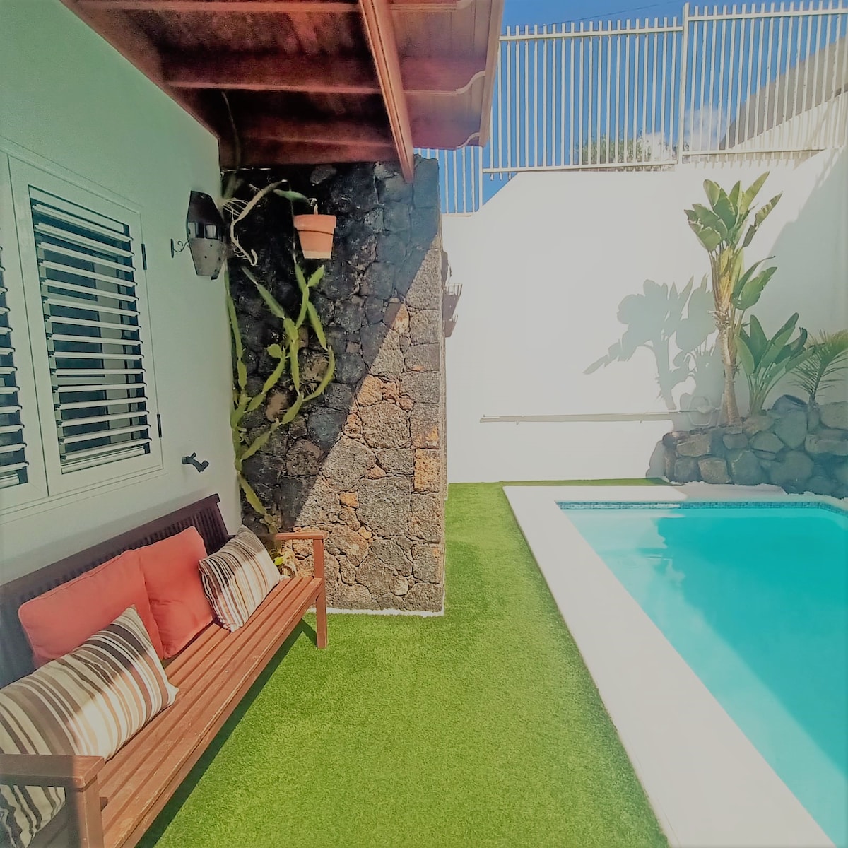 A private pool area features a well-maintained swimming pool surrounded by a green lawn. A wooden bench adorned with colorful cushions is positioned beside the pool, offering a comfortable spot to relax in the sun. The wall is decorated with natural stone and potted plants.