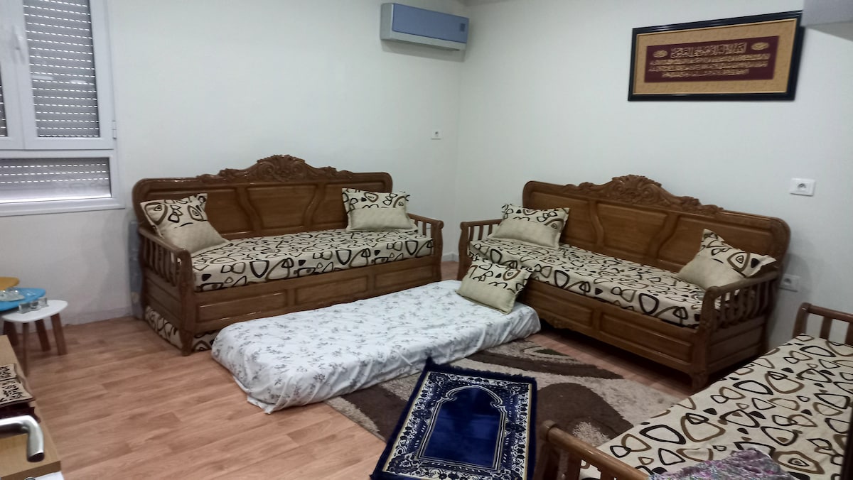A cozy living area is furnished with two wooden couches adorned with patterned cushions. A soft mat lies on the floor, complemented by a decorative rug. Natural light illuminates the space through enclosed windows, enhancing the welcoming environment.