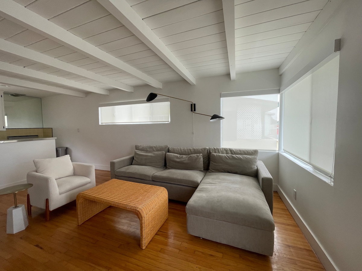 A contemporary living area features a light-colored sectional sofa accompanied by a woven coffee table. A small, white accent chair adds to the seating arrangement. Large windows allow natural light to fill the space, highlighting the white paneled ceiling.