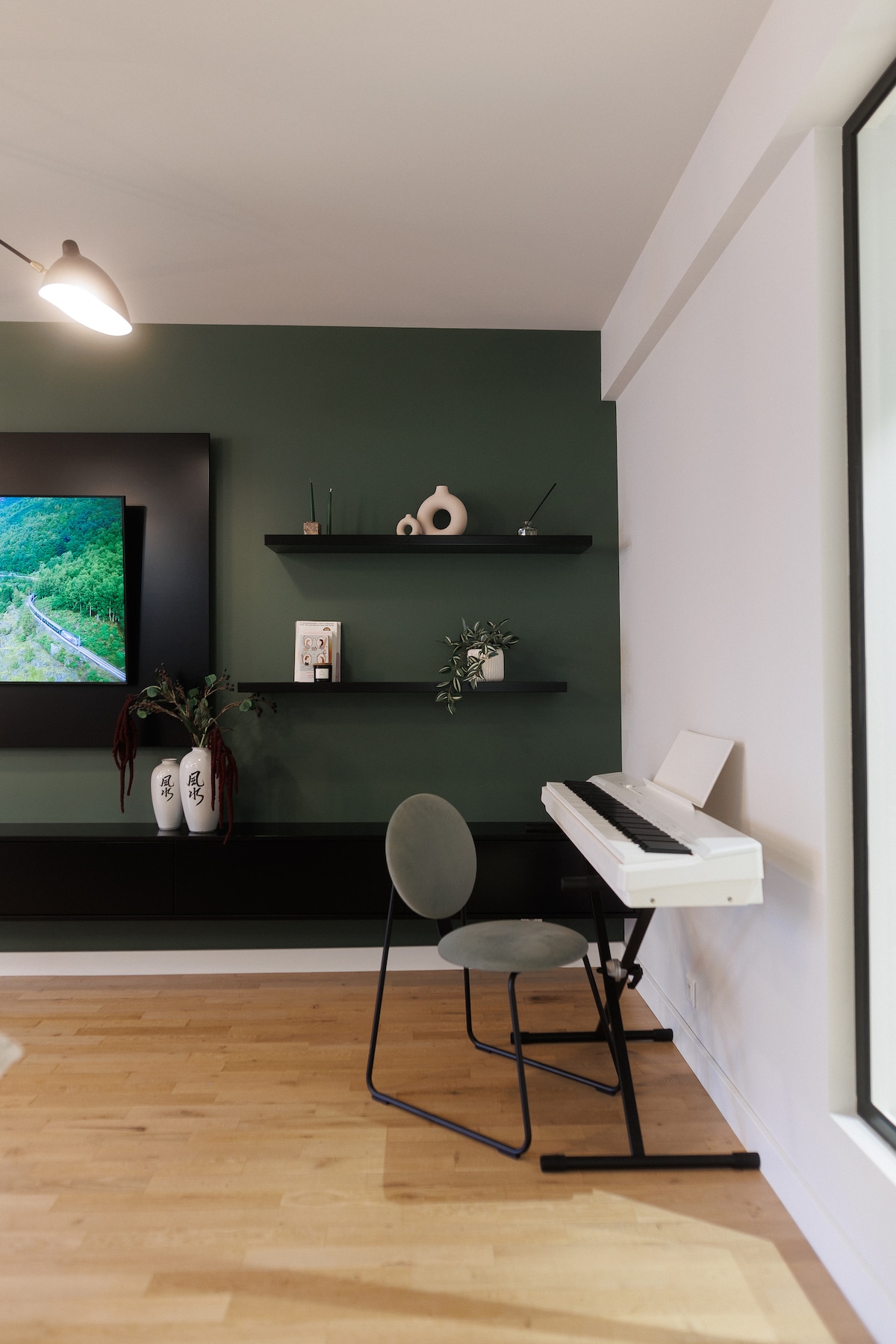 A workspace area features a digital piano alongside a minimalistic chair. Decorative shelves hold vases and small items against a deep green accent wall. A large television provides a modern touch, while light wood flooring completes the inviting atmosphere.