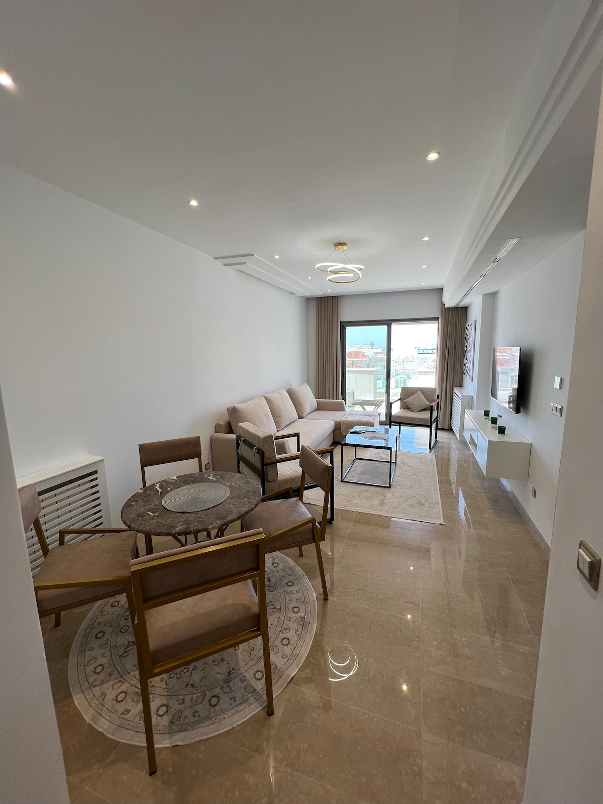 A modern living space features a light-colored sofa and armchairs arranged around a circular glass table. Natural light streams in through a sliding door that leads to a private balcony, while decorative details enhance the contemporary design.